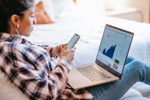 Een jonge vrouw die via haar telefoon en laptop haar investeringen aan het bekijken is.